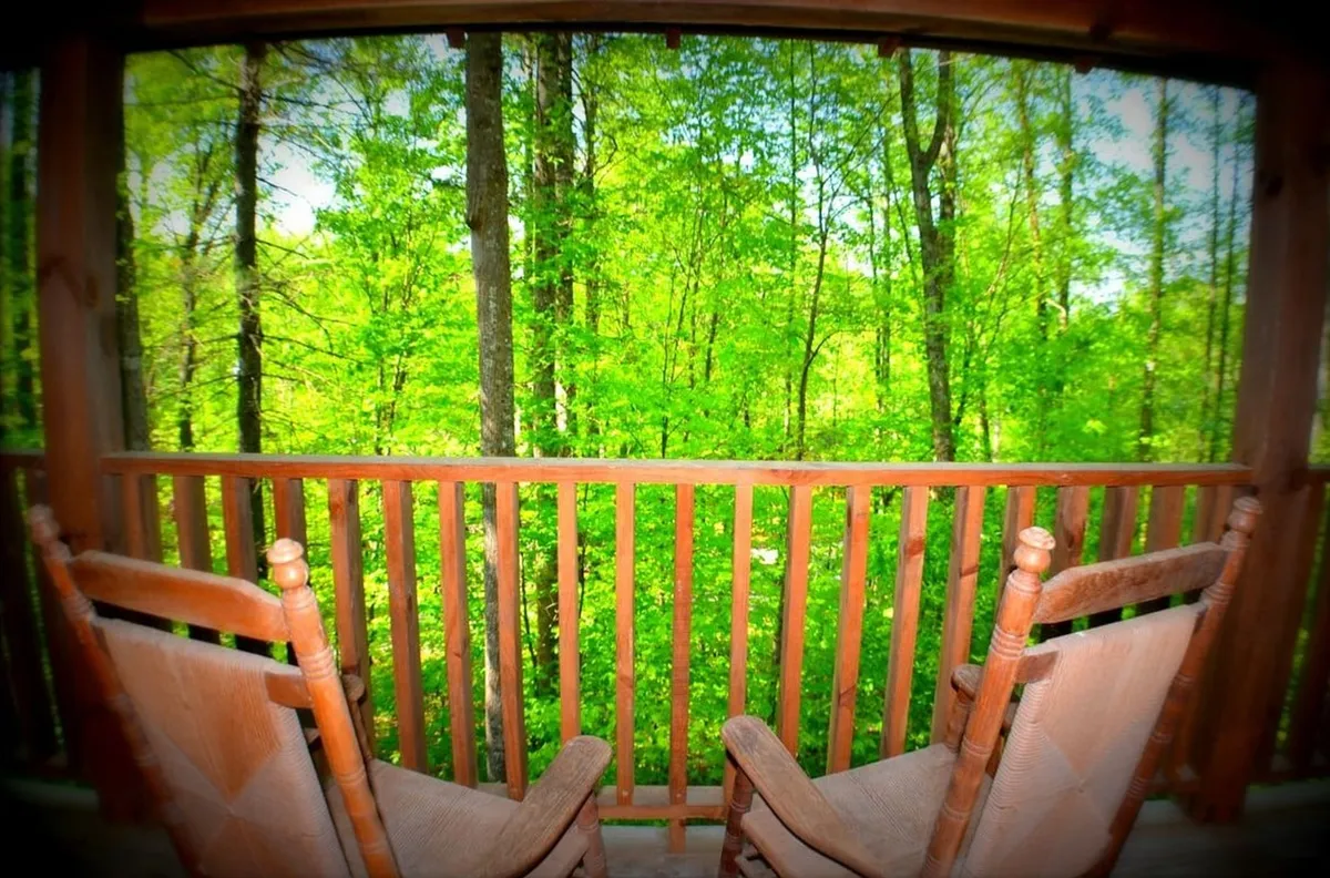 This image captures a peaceful outdoor deck scene, featuring wooden rocking chairs set against a backdrop of lush green trees and a sturdy wooden railing. The setting invites relaxation while offering expansive views of the natural forest surroundings. The mood is calm and serene, ideal for enjoying morning coffee or simply soaking up the tranquil atmosphere of the Smoky Mountains.