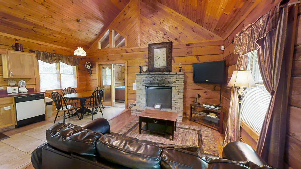 This image features a warm, inviting living room with a striking stone fireplace as its centerpiece. The space is furnished with a black leather sofa, wooden ceilings, and an adjacent dining area visible in the background. Sunlight streams through windows, enhancing the cozy and welcoming atmosphere. The room is ideal for family gatherings, relaxing by the fire, and enjoying leisure time together.