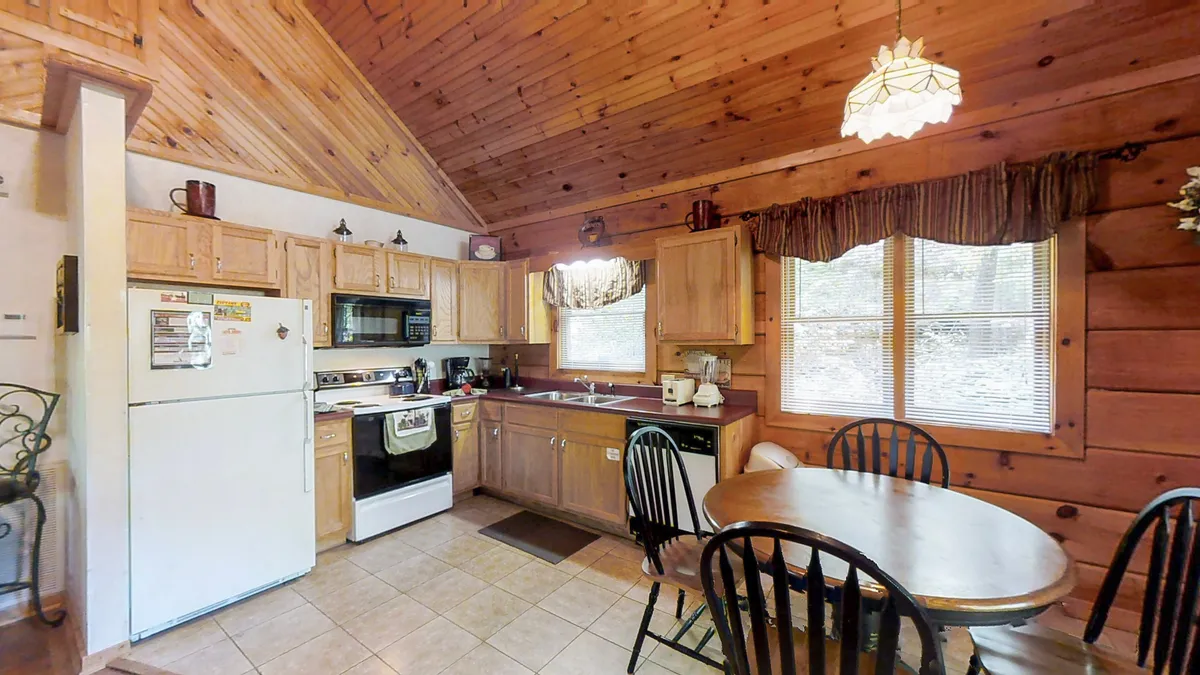 The image features a warmly lit kitchen and dining area framed by wooden cabinetry and large windows that invite natural light. A round dining table for four sits at the center, surrounded by rustic décor. The kitchen space is open and functional, equipped for cooking and socializing.